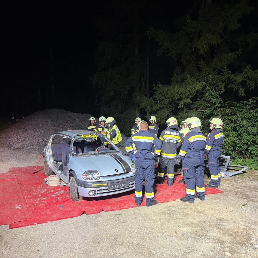 Feuerwehrleute in gelben und blauen Uniformen stehen um ein demoliertes Auto auf einem roten Teppich in der Nacht.