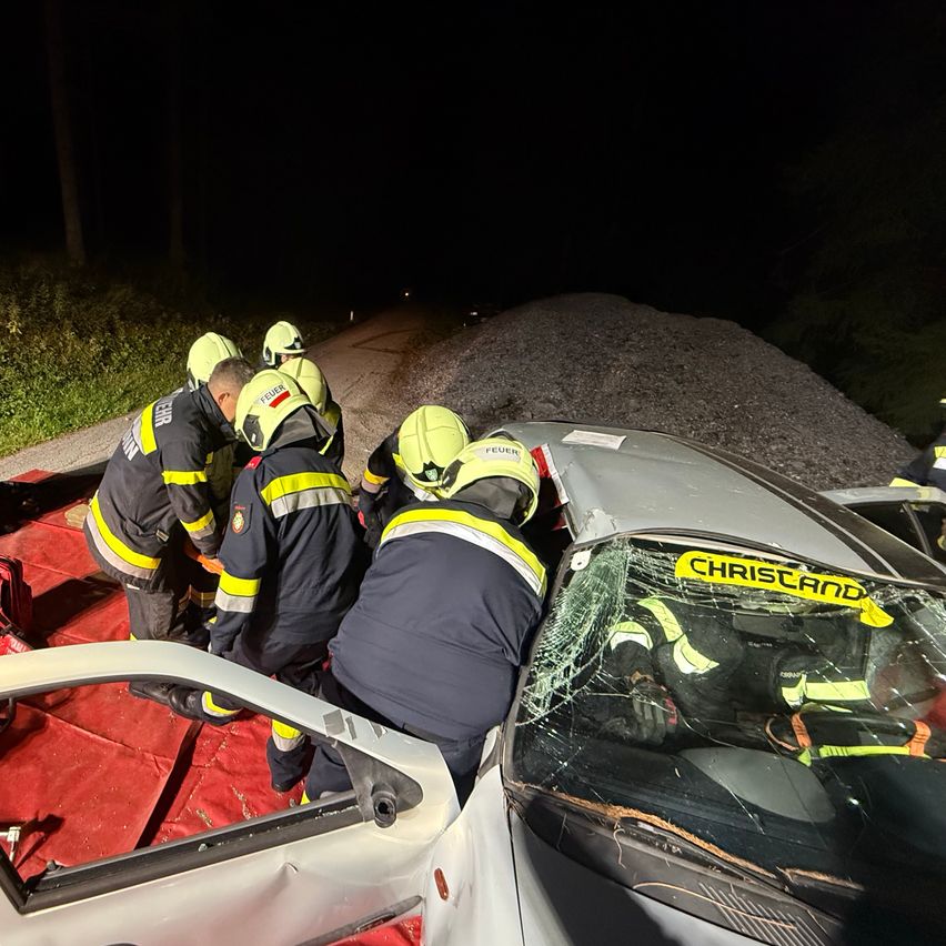Feuerwehrleute retten eine Person aus einem verunglückten Auto bei Nacht. Das Auto steht auf einem roten Tuch, und die Windschutzscheibe ist zerbrochen.