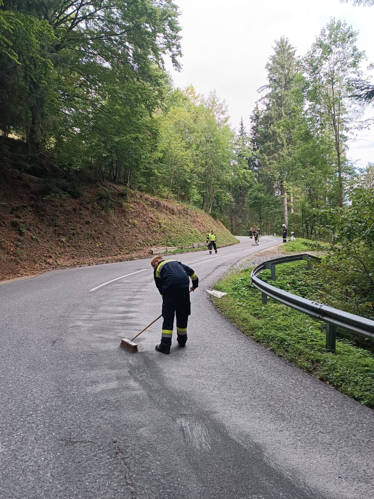 Ein Mann fegt die Straße in einem Waldgebiet, mit mehreren Personen, die im Hintergrund gehen.