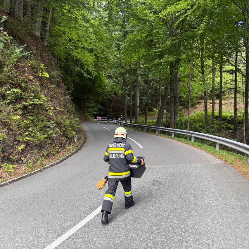 Ein Feuerwehrmann geht auf einer kurvenreichen Straße, hält ein schwarzes Objekt und trägt einen gelben und schwarzen Overall.