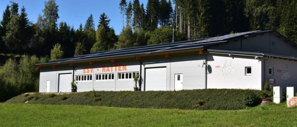 Ein weisses Gebäude mit Solarpanelen auf dem Dach, benannt ESV Ratten, ist von grünem Gras und Bäumen umgeben. Es gibt zwei grosse weisse Türen und eine kleinere weisse Tür.