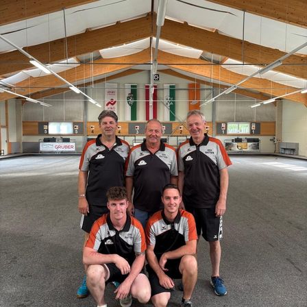 Fünf Männer in Sportuniformen posieren für ein Foto in einer Indoor-Sportanlage mit Holzbalken und Bannern an der Wand.