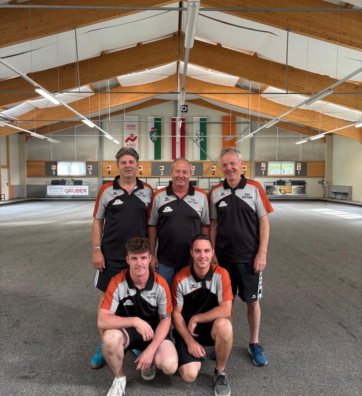 Fünf Männer in Sportuniformen posieren für ein Foto in einer Indoor-Sportanlage mit Holzbalken und Bannern an der Wand.