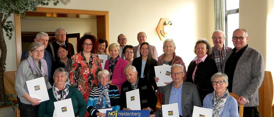 Eine Gruppe älterer Personen ist für eine Versammlung in einem Raum versammelt. Sie halten Papiere mit dem Wort NEIN darauf.