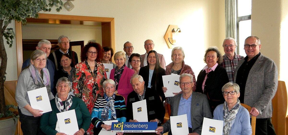 Eine Gruppe älterer Personen ist für eine Versammlung in einem Raum versammelt. Sie halten Papiere mit dem Wort NEIN darauf.