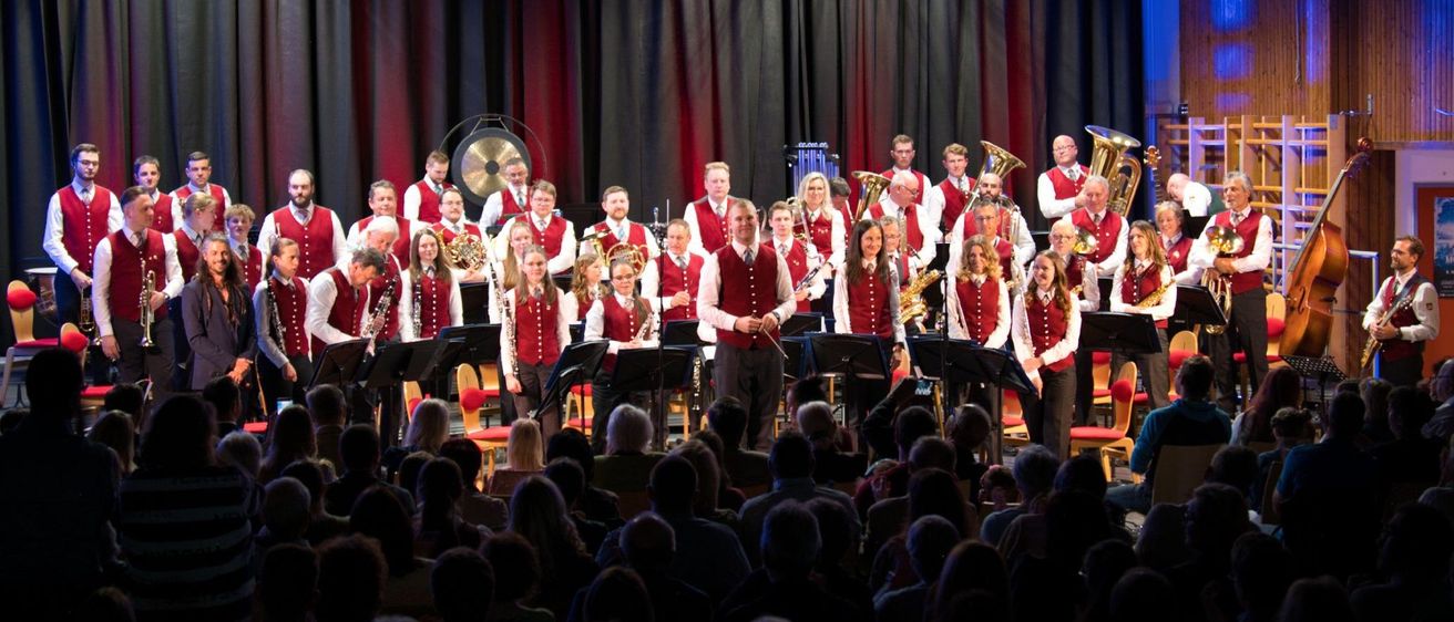 Eine Musikergruppe spielt auf der Bühne mit Instrumenten wie Tubas und Trompeten und trägt passende rote Westen. Dahinter befinden sich rote und schwarze Vorhänge. Das Publikum sitzt in Stühlen vor ihnen.