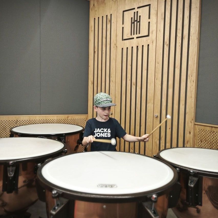 Ein junger Junge in einem Jack & Jones T-Shirt und einer Camouflage-Mütze spielt in einem Raum mit Holzwänden Trommeln.