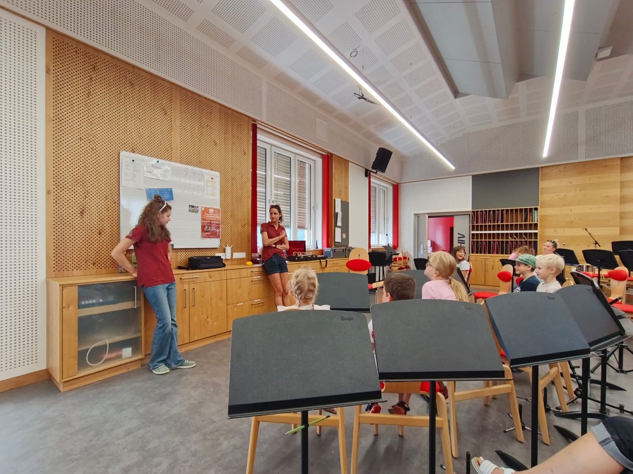 Ein Klassenzimmer mit Holzwänden und einer Tafel, wo eine Frau neben einem Schreibtisch steht und eine andere Frau vor einer Gruppe von Kindern steht, die auf Stühlen sitzen.