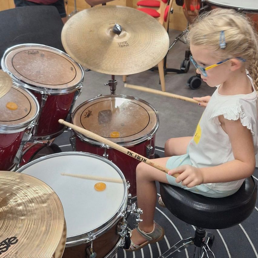 Ein junges Mädchen mit Brille und geflochtener Frisur sitzt auf einem Hocker und spielt Schlagzeug. Sie hält Drumsticks in den Händen und um sie herum stehen Schlagzeuge und Becken.