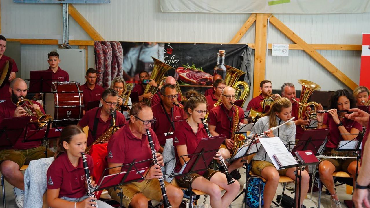 Eine Gruppe von Musikern in roten Hemden und mit Brillen spielt verschiedene Instrumente, darunter Klarinetten, Saxophone und Trompeten, in einem Raum mit Holzbalken und einem Banner im Hintergrund.