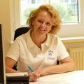 Eine Frau mit blonden Haaren lächelt in die Kamera, während sie an einem Schreibtisch sitzt, einen Stift hält und ein weißes Polo-Shirt mit dem Text 'KIR SE' trägt. Hinter ihr befindet sich ein Fenster mit Blick auf Bäume.