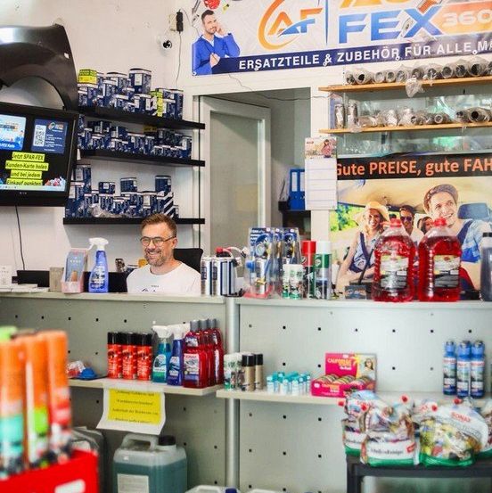 Ein Mann steht hinter einem Tresen in einem Geschäft mit verschiedenen Produkten und einem Monitor. Der Laden ist gut bestückt mit Reinigungsmitteln, Flüssigkeiten und Flaschen.