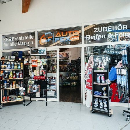 Ein Ladeneingang mit einer Glastür, Regalen voller Produkte und einer Glaswand, auf der 'AUTO FEX 360' und 'KFZ Ersatzteile für alle Marken' steht. Im Inneren des Ladens sind weitere Produkte ausgestellt.