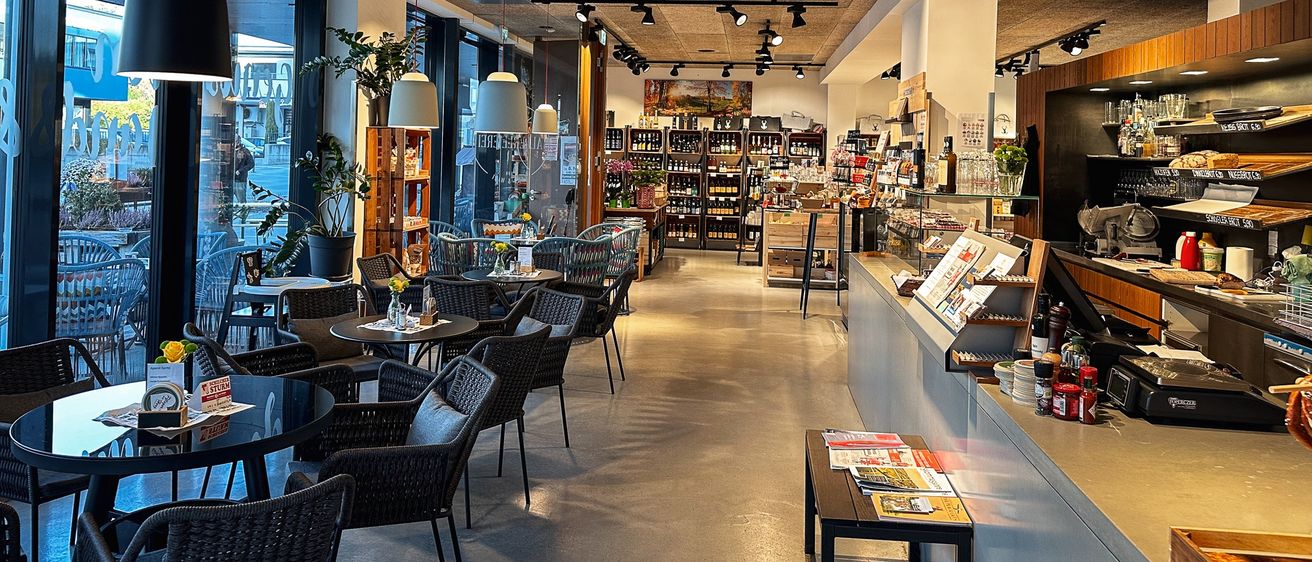 Das Innere eines modernen Cafés mit großen Fenstern, Tischen und Regalen mit Flaschen. Es hat einen langen Tresen mit Zeitschriften und Gewürzen.