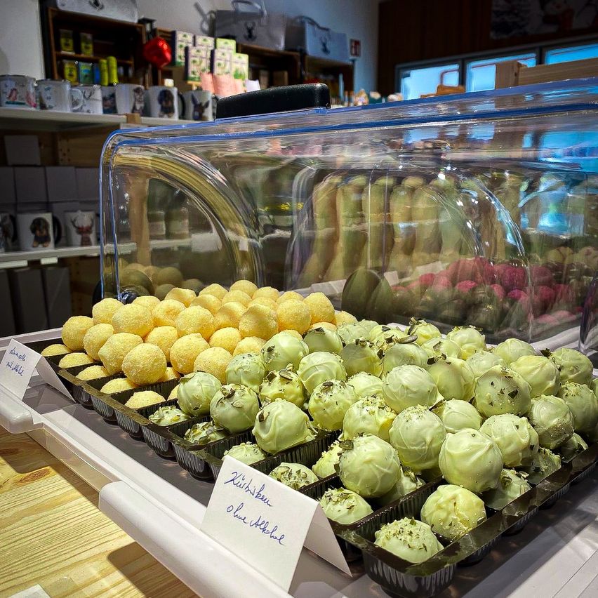 Eine Vitrine mit verschiedenen Pralinen, darunter gelbe und grüne Sorten, auf einem Holztisch in einem Geschäft angeordnet. Im Hintergrund befinden sich Regale mit verschiedenen Artikeln.