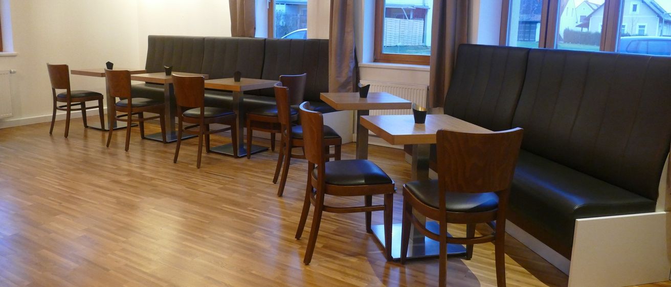 Ein leerer Essbereich mit Holzböden, Lederstühlen und runden Tischen. Bankreihen sind an der Wand mit Fenstern aufgestellt. Ein Bilderrahmen befindet sich an der Wand über den Fenstern.