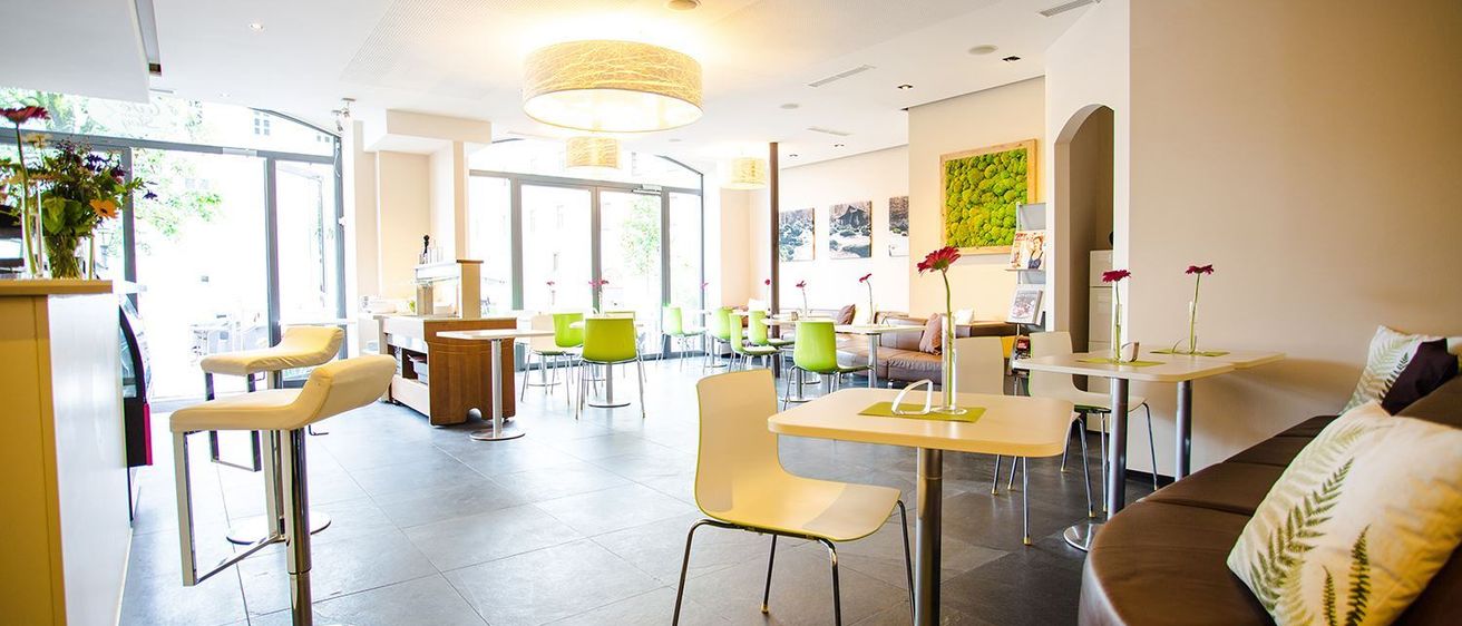 Ein leeres modernes Café mit weißen und grünen Stühlen um runde Tische, einem großen Fenster links und dekorativer Kunst an der Wand.