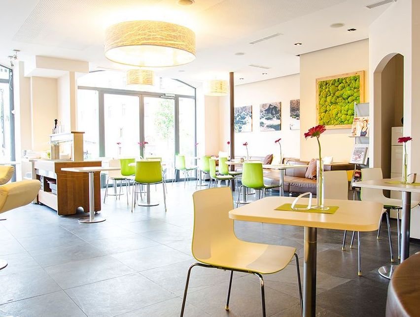 Ein leeres modernes Café mit weißen und grünen Stühlen um runde Tische, einem großen Fenster links und dekorativer Kunst an der Wand.