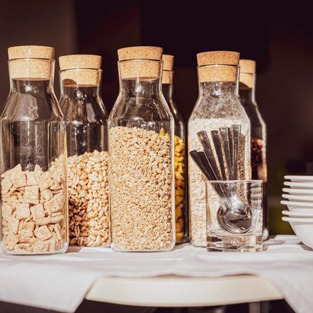 Ein Tisch zeigt Glasflaschen mit verschiedenen Cerealien und Hafer, verschlossen mit Korkstopfen. Ein Glas mit Löffeln steht daneben, und weiße Teller und Schüsseln sind rechts angeordnet.