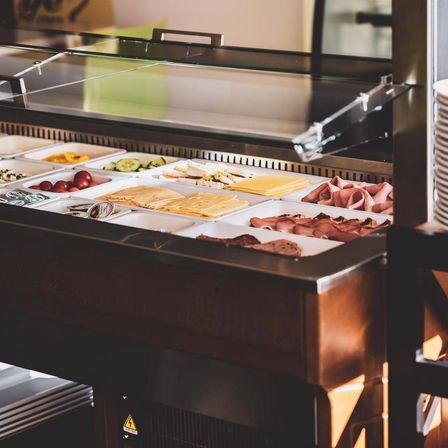 Ein Buffet-Display mit verschiedenen Speisen unter einer Glasabdeckung. Es gibt mehrere Tabletts mit Käse, Fleisch und Gemüse.