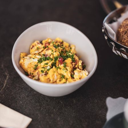 Ein weißer Teller mit Rührei, belegt mit grünen Zwiebeln und Speck, auf einer schwarzen Oberfläche.