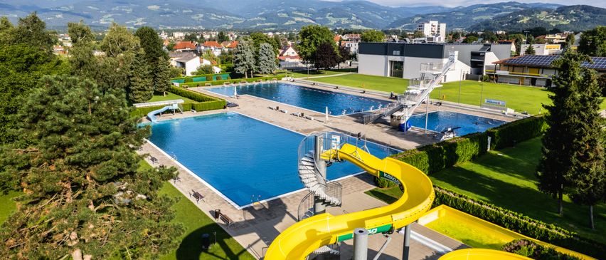 Ein Luftbild eines großen Freibades mit einer gelben Wasserrutsche, umgeben von üppigem grünem Rasen, Bäumen und Gebäuden. Der Pool ist groß und rechteckig, mit zwei Bahnen für Schwimmer.