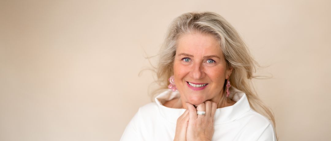 Eine lächelnde Frau mittleren Alters mit blondem Haar trägt ein weißes Hemd, rosa Ohrringe und eine Uhr.