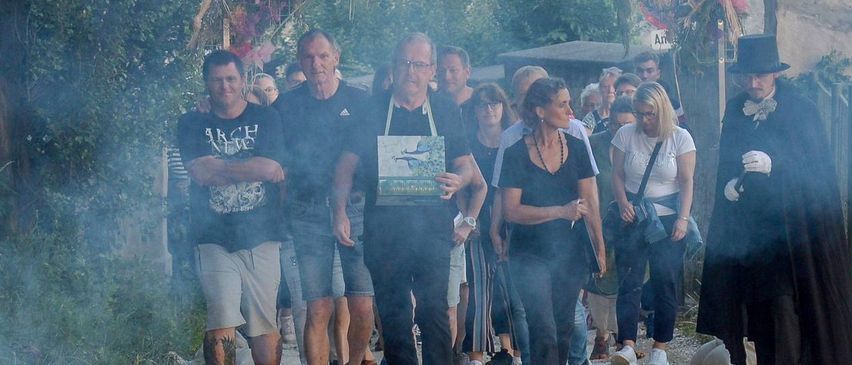 Eine Gruppe von Menschen geht auf einem von Bäumen umgebenen Pfad, mit Rauch und einem Mann in einem schwarzen Hut, der sie anführt.