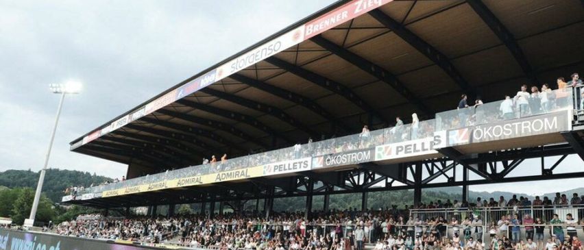 Ein Stadion gefüllt mit Zuschauern unter einem bewölkten Himmel, mit mehreren Werbebannern, darunter Admiral, Pellis und Okostrom. Einige Leute stehen auf den Geländern der oberen Tribüne.