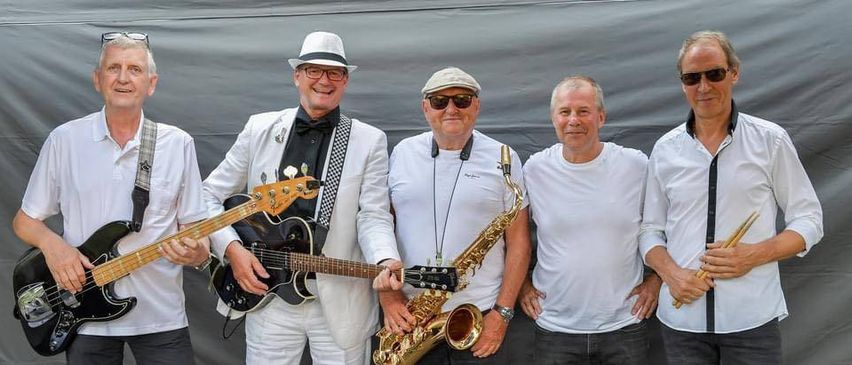 Fünf Musiker posieren für ein Foto, einer spielt Gitarre, ein anderer Saxophon, die anderen lächeln. Sie tragen weiße Hemden und Hüte. Der Hintergrund ist eine graue Plane.