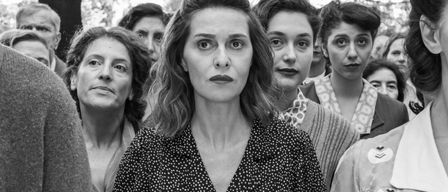 Eine Gruppe von Frauen in schwarz-weißer Kleidung steht in einem Außenbereich eng beieinander. Die Frau in der Mitte hält ein kleines Objekt in ihren Händen.