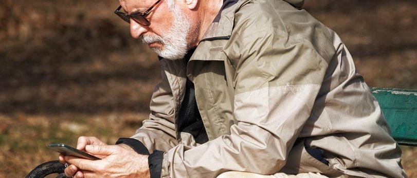Ein älterer Mann mit weißem Bart und Brille, der eine beige Jacke trägt, sitzt auf einer grünen Bank und schaut auf sein Smartphone.