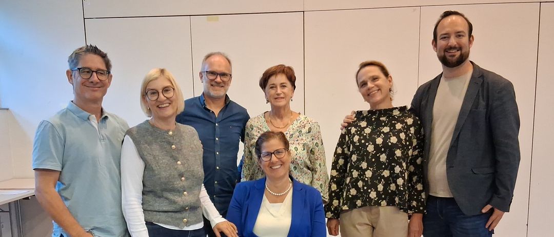 A group of seven adults stands indoors in a room with wooden floors. They are smiling and appear to be posing for a photograph. There are two women sitting on a chair.