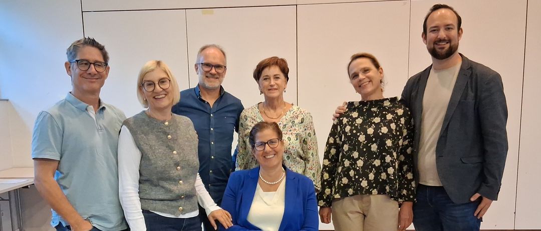 A group of six adults, including two women, stand and pose for a photo in a room with wooden floors and white walls.