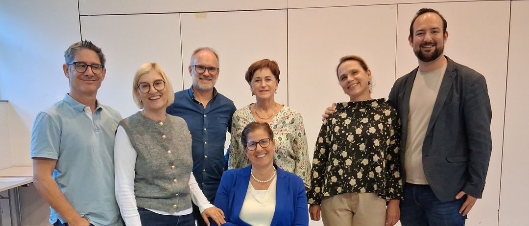 A group of seven people, including a woman in blue sitting on a chair, pose for a photo on a wooden floor.