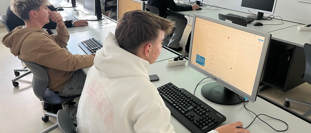 Mehrere Schüler sitzen in einem Klassenzimmer und benutzen Computer. Ein Schüler trägt eine weiße Kapuzenjacke und benutzt eine Maus. Ein anderer Schüler trägt eine braune Jacke und hat die Hand im Kinn.