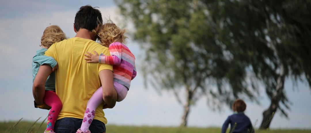 Ein Mann trägt zwei kleine Kinder auf seinen Schultern in einem Feld voller roter Blumen. Ein Junge im Hintergrund geht auf sie zu.