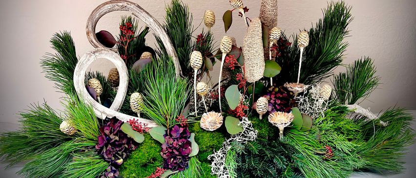 Eine dekorative Blumenarrangement besteht aus verschiedenen grünen Blättern, Moos und lila Blumen, mit hohen, strukturierten Elementen und dekorativen weißen Geweihen.