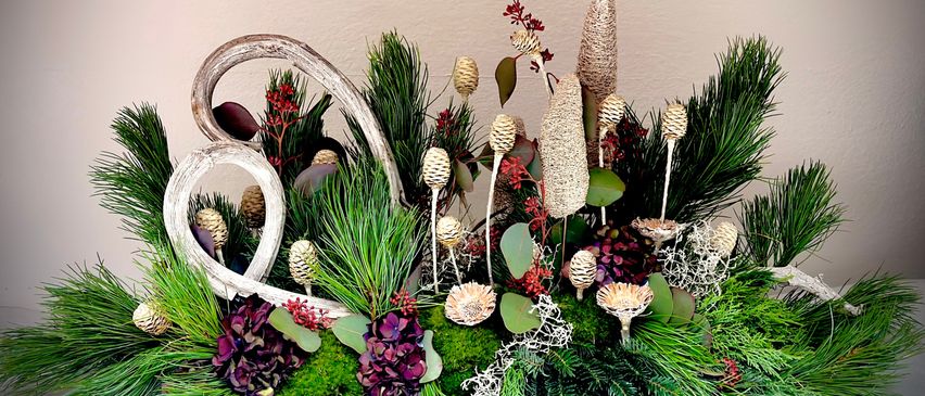 Eine künstlerische Blumenarrangement mit einer Mischung aus Grünpflanzen, lila Blumen und dekorativen Elementen wie Holzzweigen und Zapfen auf einer weißen Oberfläche.