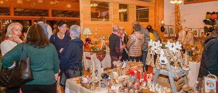 A group of people gather around a table filled with Christmas decorations and ornaments. The room is warmly lit with wooden walls and ceiling lamps.