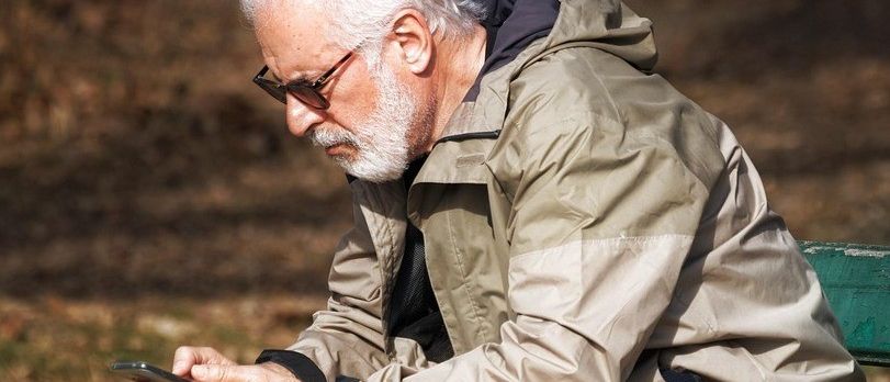 Ein älterer Mann sitzt auf einer grünen Parkbank. Er trägt eine Brille, eine beige Jacke und beige Hosen. Er benutzt ein Smartphone.