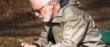 Ein älterer Mann sitzt auf einer grünen Parkbank. Er trägt eine Brille, eine beige Jacke und beige Hosen. Er benutzt ein Smartphone.