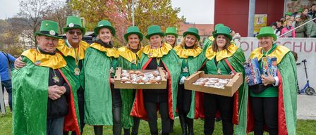 Eine Gruppe von Personen, die grüne und gelbe Kostüme tragen und Schachteln mit Donuts halten, steht auf einem Rasenfeld. Hinter ihnen befinden sich Bäume und ein rotes Gebäude.