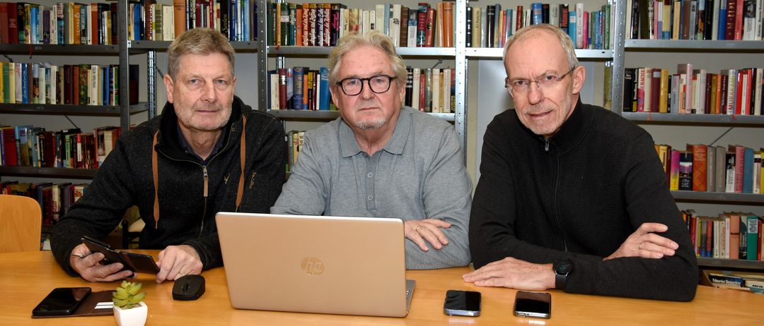 Drei Männer in Freizeitkleidung sitzen um einen Tisch in einer Bibliothek, lächeln und posieren für ein Foto. Ein Laptop, Handys und eine kleine Topfpflanze stehen auf dem Tisch. Dahinter sind mit Büchern gefüllte Regale zu sehen.