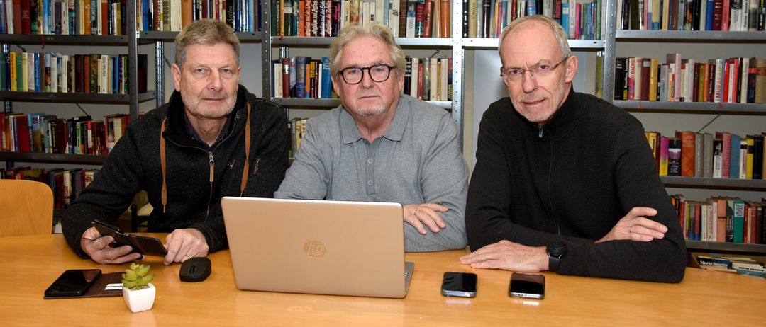 Drei Männer in einer Bibliothek, einer mit Brille, einer mit Laptop und der dritte mit Smartphone.