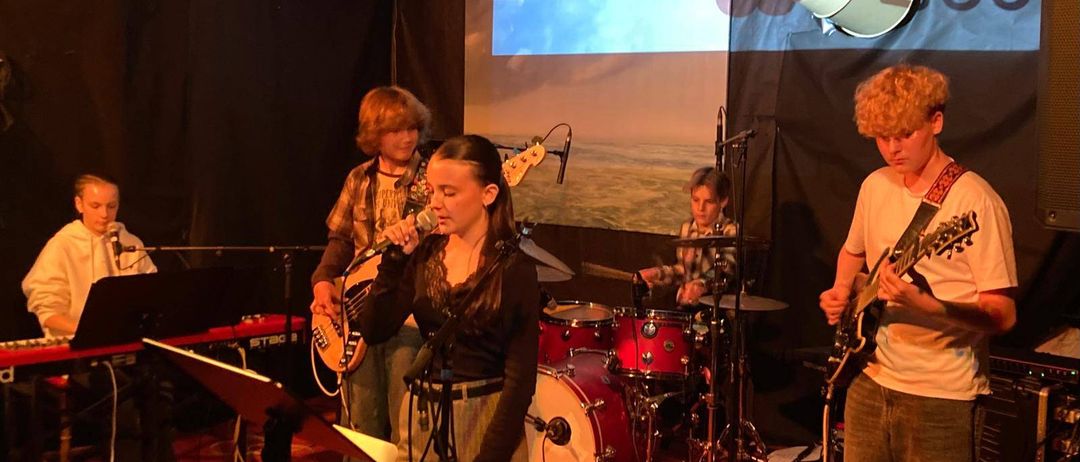 Eine Band tritt auf der Bühne auf, vor einem Hintergrund mit Sonnenuntergang. Die Sängerin, eine Frau, hält ein Mikrofon. Ein Junge spielt Schlagzeug, ein anderer Gitarre. Die Bühne ist mit verschiedenen Instrumenten ausgestattet.