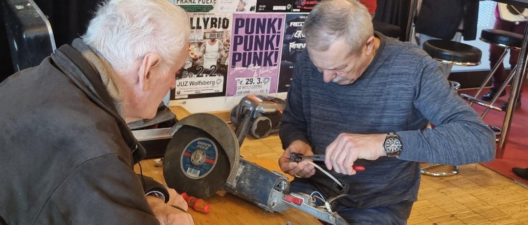 Zwei ältere Männer arbeiten an einer Maschine mit einem Kreissägenblatt. Einer hält einen Draht, während der andere ein Werkzeug benutzt. Sie sind in einem Raum mit einem Holzboden und Postern an der Wand.