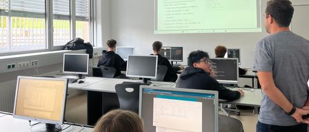 Ein Klassenzimmer mit Computerbildschirmen, Schülern und einem Lehrer. Die Projektionsleinwand zeigt eine Webseite über div-Karten.