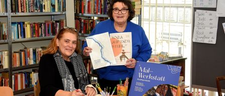 Zwei Frauen sitzen an einem Tisch in einer Bibliothek. Eine Frau hält ein Buch mit dem Titel 'Yoga auf dem Stuhl', während die andere ein Notizbuch hält und zeichnet. Sie sind umgeben von Büchern und einem Schild mit 'Mal-Werkstatt' darauf.