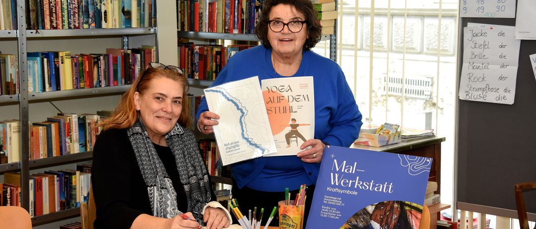 Zwei Frauen sitzen an einem Tisch in einer Bibliothek. Eine Frau hält ein Buch mit dem Titel 'Yoga auf dem Stuhl', während die andere ein Notizbuch hält und zeichnet. Sie sind umgeben von Büchern und einem Schild mit 'Mal-Werkstatt' darauf.